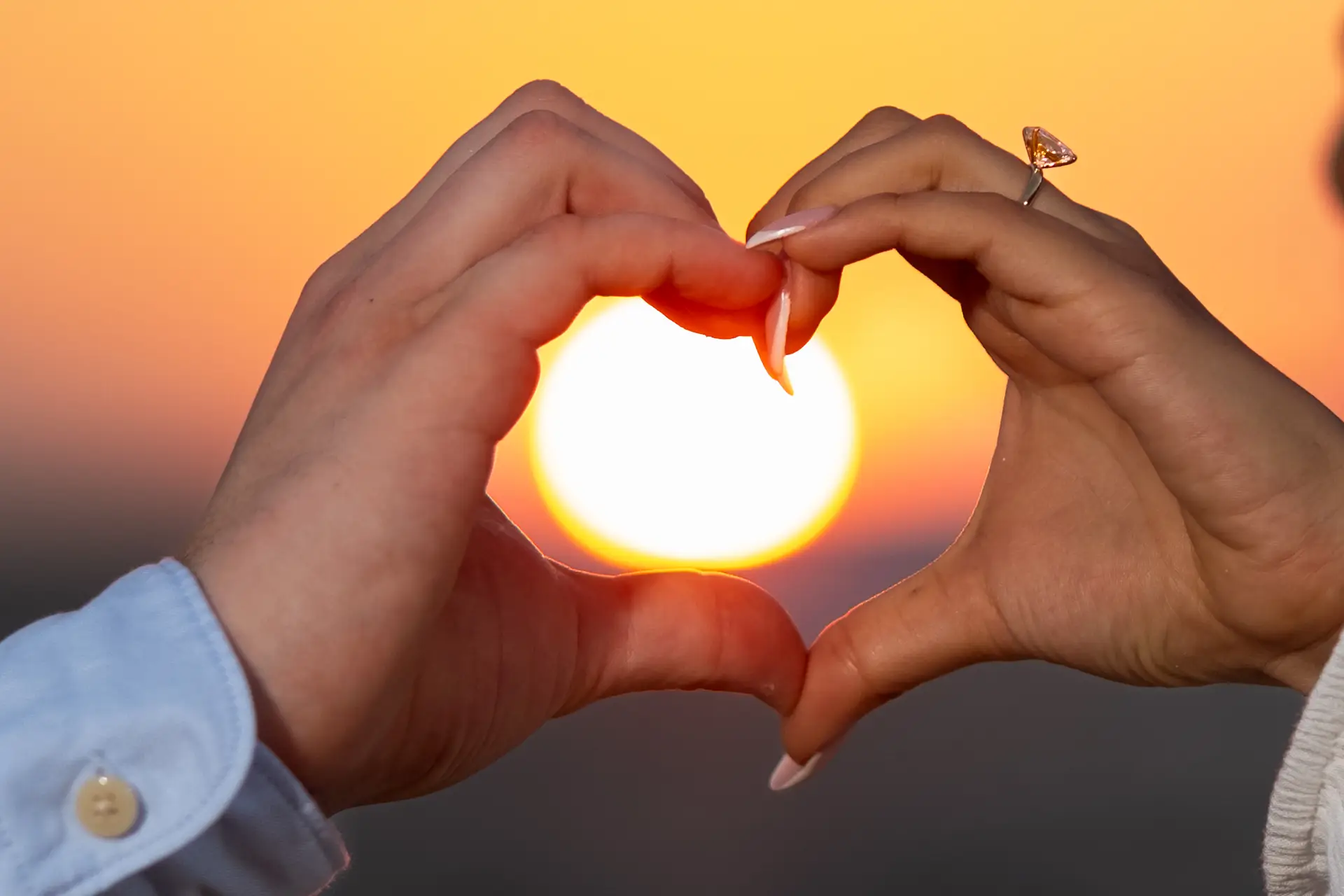 Couple forming a heart with their hands around the sunset, showing their engagement ring