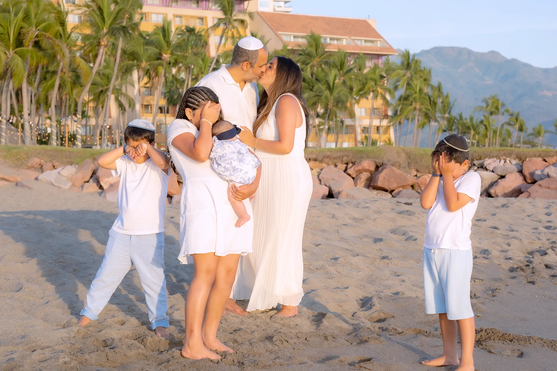 Parents kissing while holding their baby as their children play on the beach in Puerto Vallarta