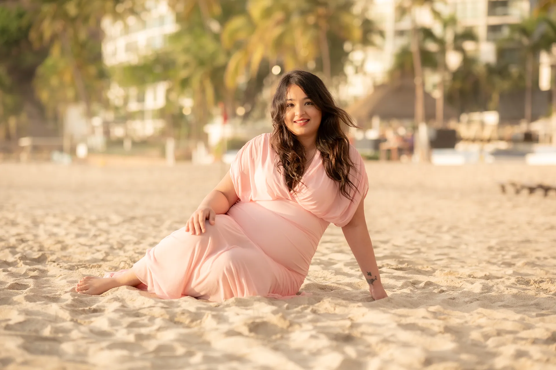 Expecting mother in a pink dress seated on the sand during a maternity session in Puerto Vallarta