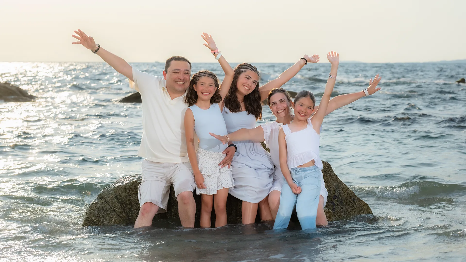 Family of five with arms raised, seated on a rock in the shallows at sunset in Puerto Vallarta
